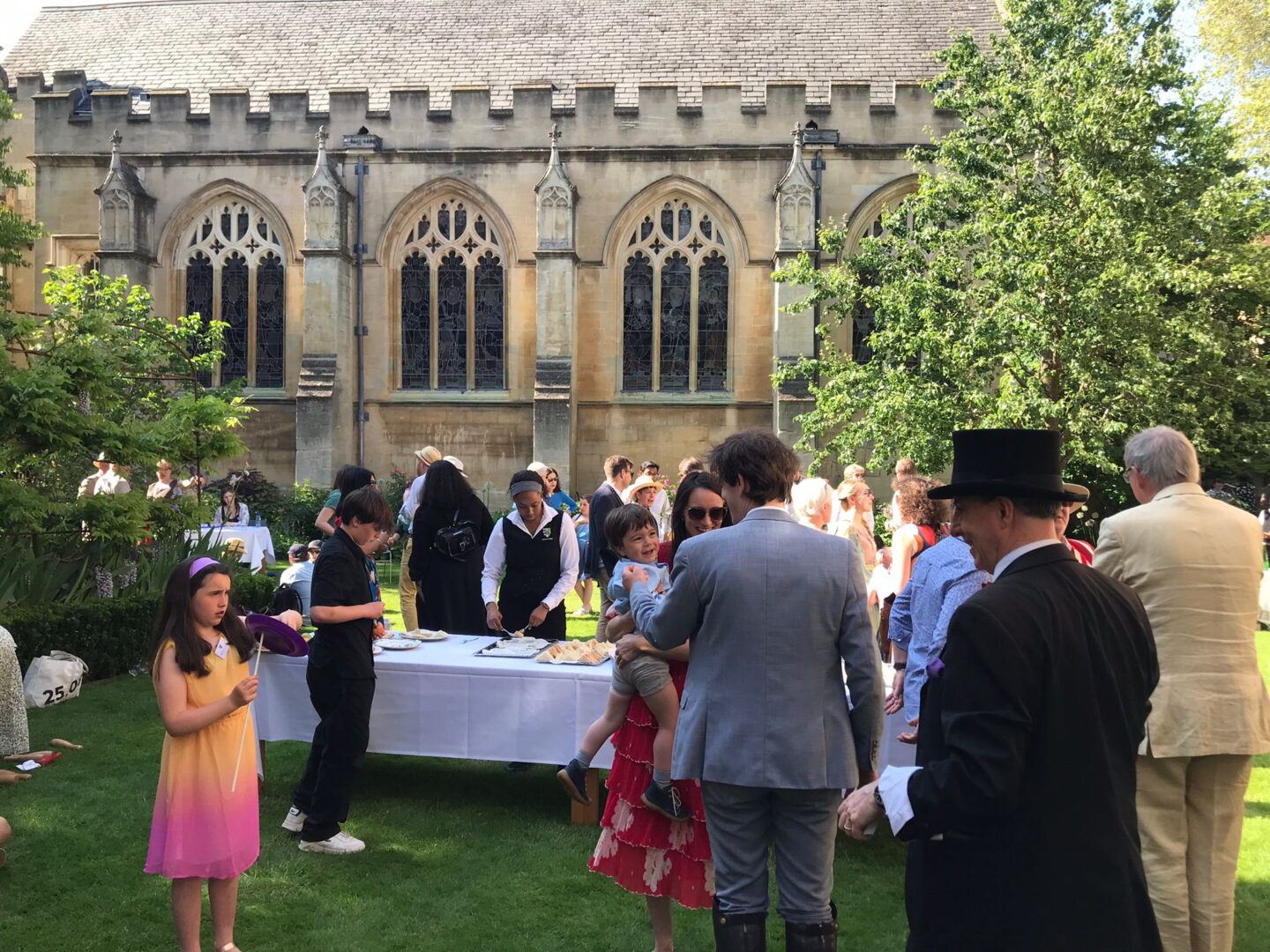 Photo of garden party in Rector's Garden with man in top hat in foreground, a child in an orange and pink dress to the left, and other people milling around a table covered in a white table cloth