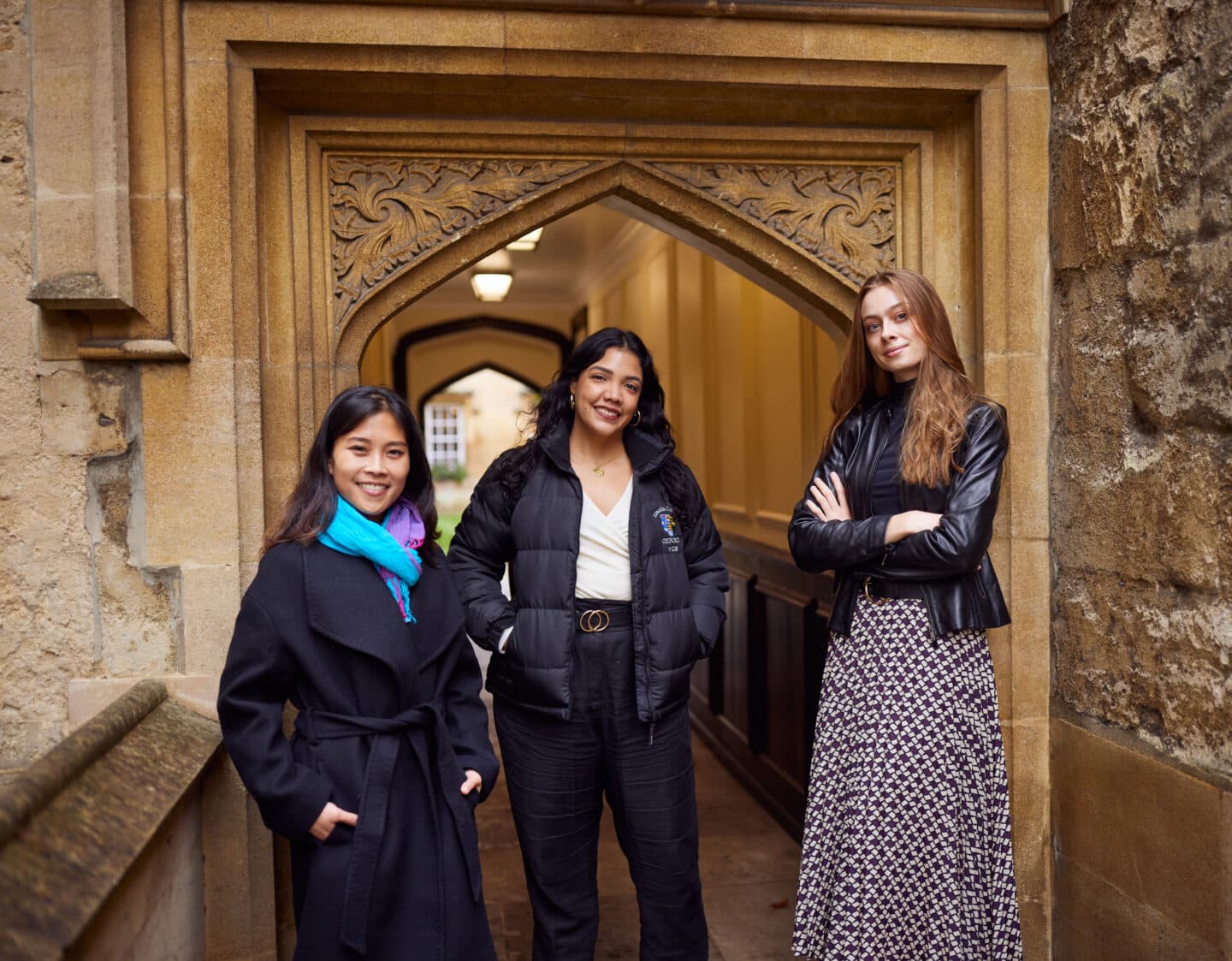 Three female students standing in front of an old arched doorway in Lincoln College, smiling at the camera