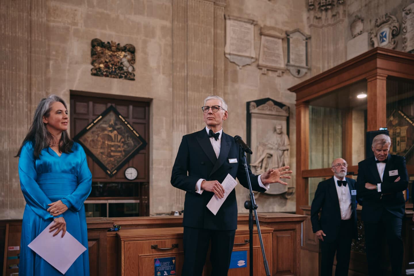 The Rector in a black tuxedo speaks at a microphone inside Lincoln College Library, with other guests and Lucy Matheson in a blue dress standing nearby against a backdrop of historic stonework and memorial plaques.