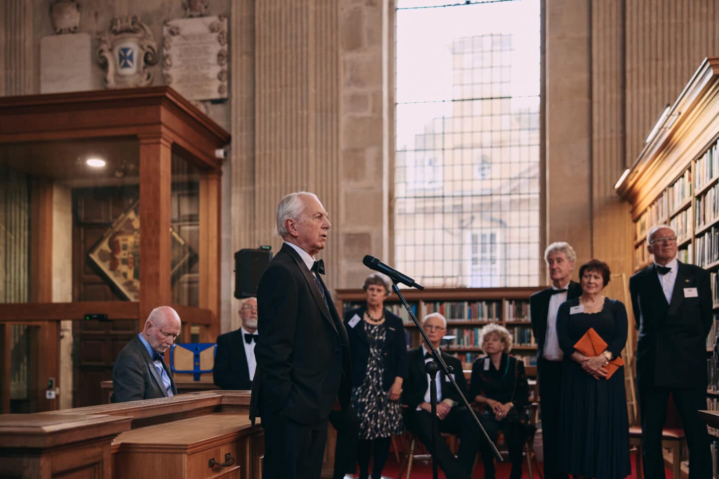Supernumerary Fellow, Nigel Wilson, in a tuxedo addresses attendees from a microphone in Lincoln College Library, while a group of formally dressed guests listen attentively among the bookshelves.