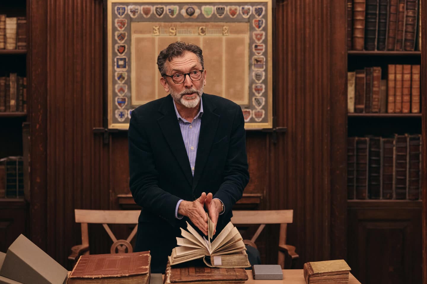 Fellow Peter McCullough with glasses and a beard shows an old book during a presentation in Lincoln College Library, surrounded by antique volumes and a decorative shield display on the wall behind him.
