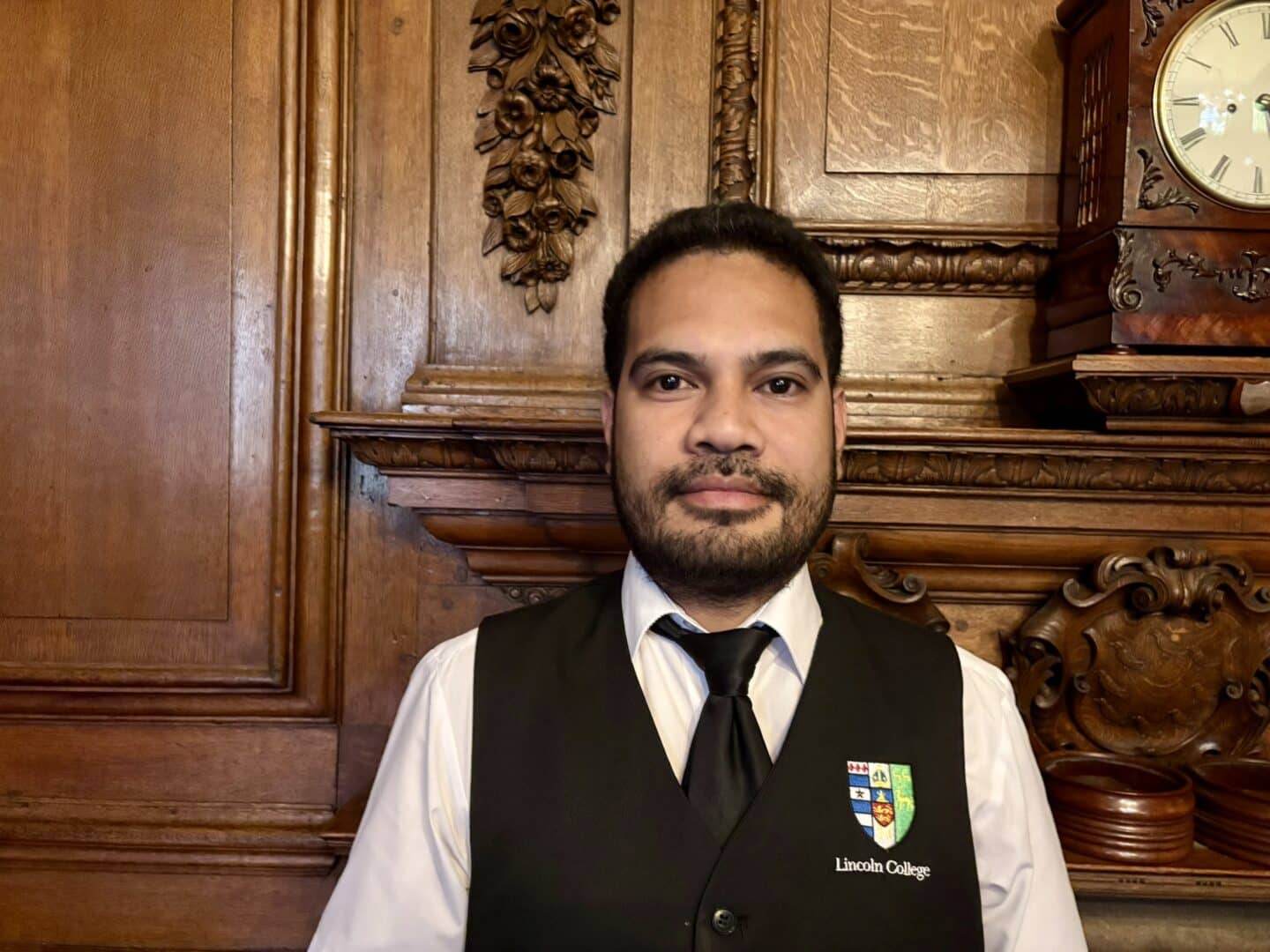 Adelino De Araujo Mendonca, Catering Supervisor at Lincoln College, standing in front of an ornate wooden fireplace with carved detailing and a clock above, wearing a white shirt, black waistcoat, and tie with the College crest.