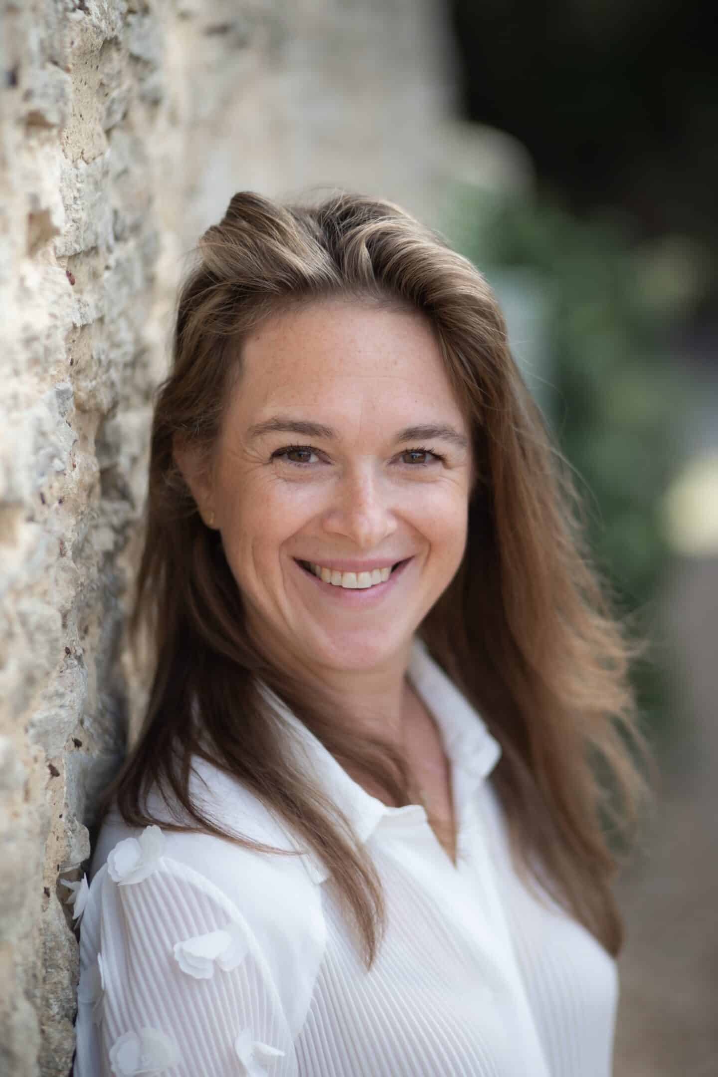 Portrait of Arabella Begin smiling outdoors, leaning against a stone wall, wearing a white blouse.