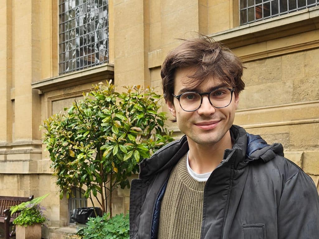 Artur Harris with brown hair and round glasses smiles at the camera while standing outside a historic stone building with leaded windows at Lincoln College. He wears a dark jacket over a khaki jumper and white T-shirt. A leafy shrub and wooden bench sit in the background.