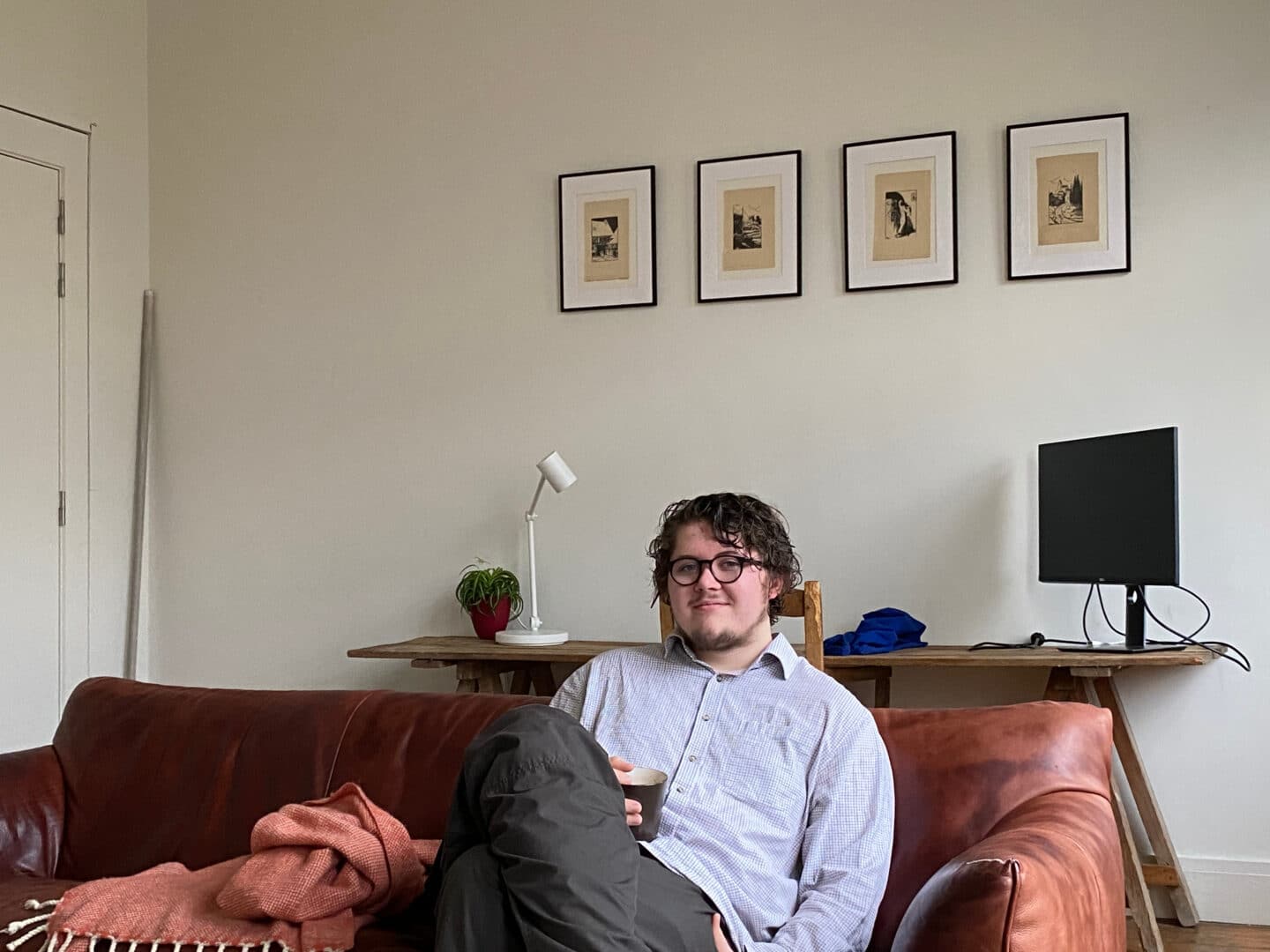 Austin Spendlowe seated on a brown leather sofa, holding a mug and smiling towards the camera. Behind him, a minimalist workspace with framed prints, a houseplant, and a monitor adds a calm, homely atmosphere.