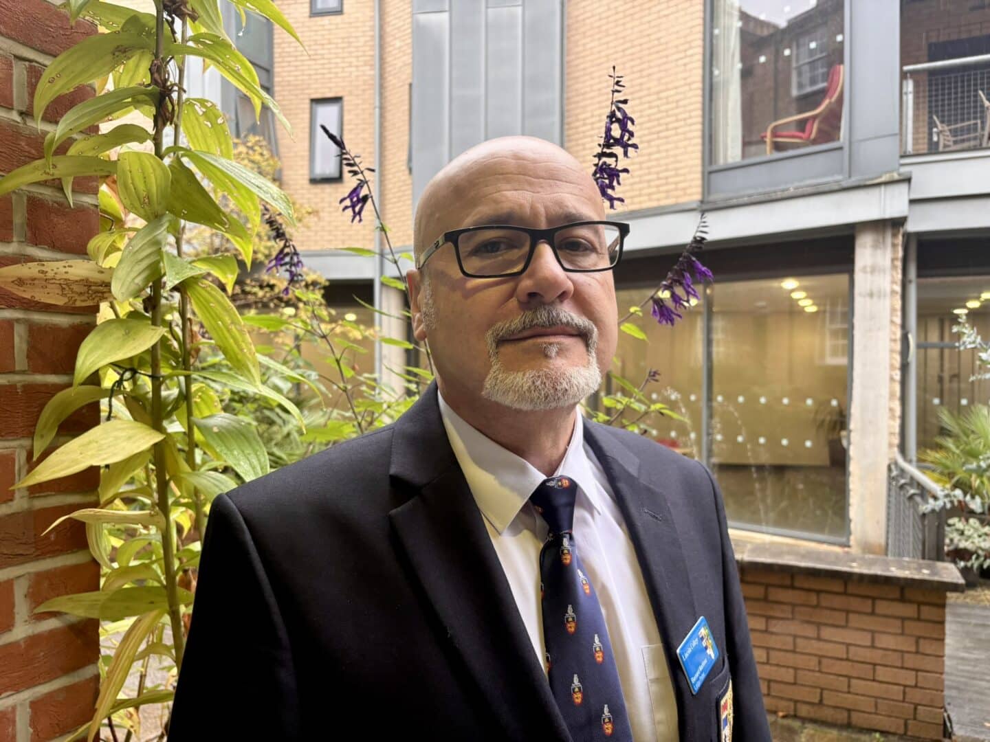 Bernard Martinez, a Lincoln College porter, standing in a courtyard beside a brick pillar and flowering plants, wearing a dark suit, tie, and name badge, with College buildings in the background.