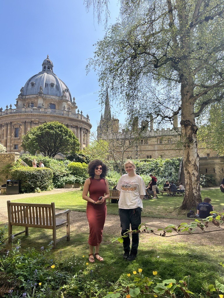 Two Lincoln students standing in the University Parks with the Radcliffe Camera and Lincoln College library tower in the background, taking part in the Green Impact Bioblitz biodiversity survey on a sunny day.
