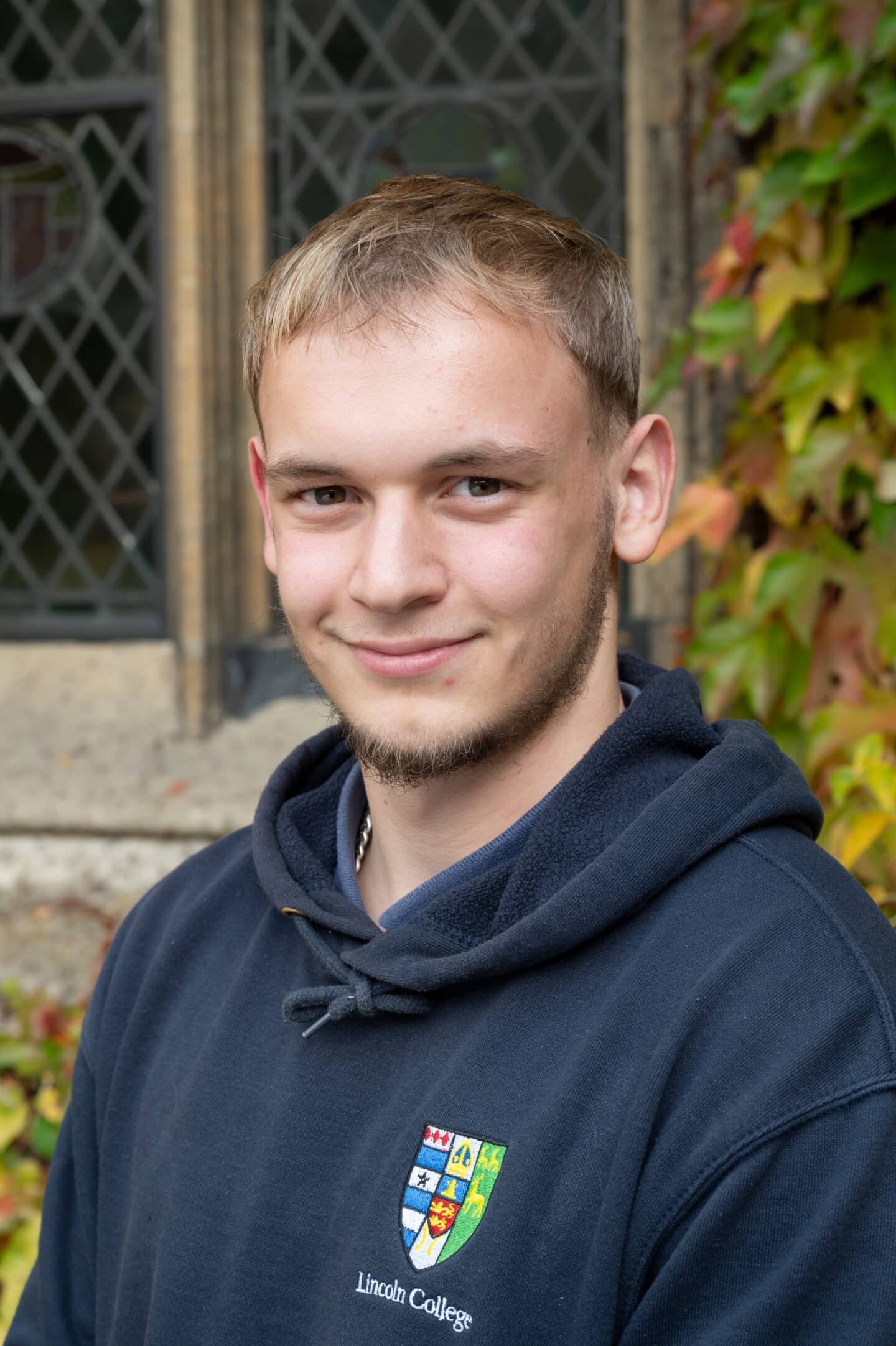 Harry Pearce standing in Front Quad at Lincoln College, Oxford, wearing a navy Lincoln College hoodie, with ivy and a leaded window behind