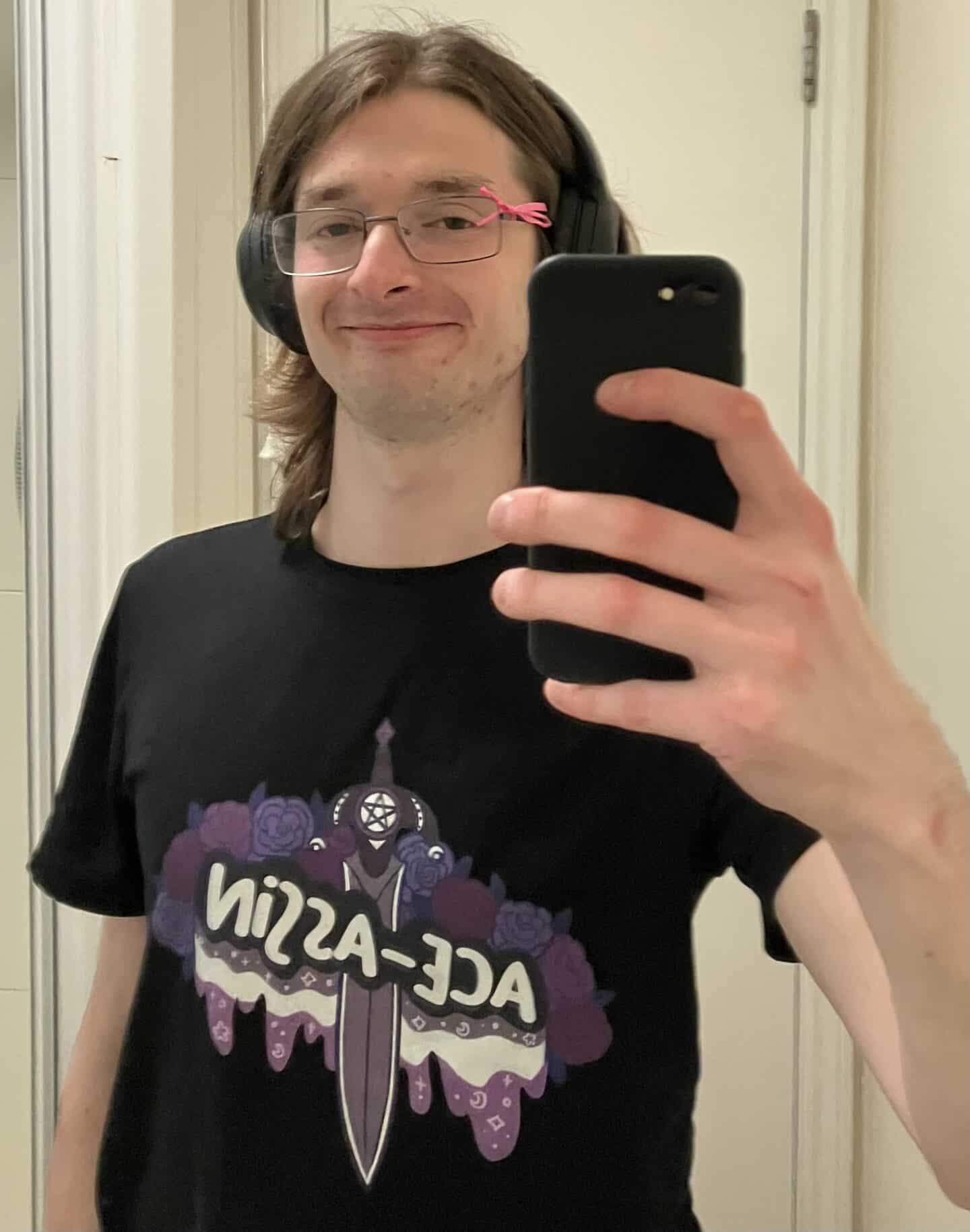 Charlie Franklin smiling in a mirror selfie, wearing headphones and a black T-shirt with a bold graphic design that reads 'ACE-ASSIN' over a purple sword and roses illustration.