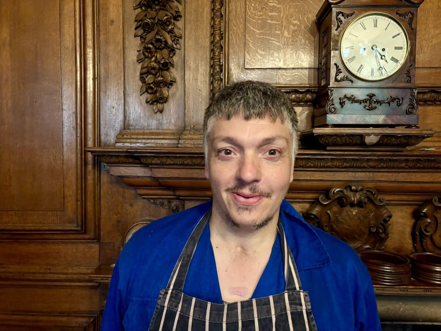“Christopher Ray, a Lincoln College kitchen porter, standing in front of an ornate wooden fireplace with carved detailing and a clock above, wearing a blue uniform and striped apron.