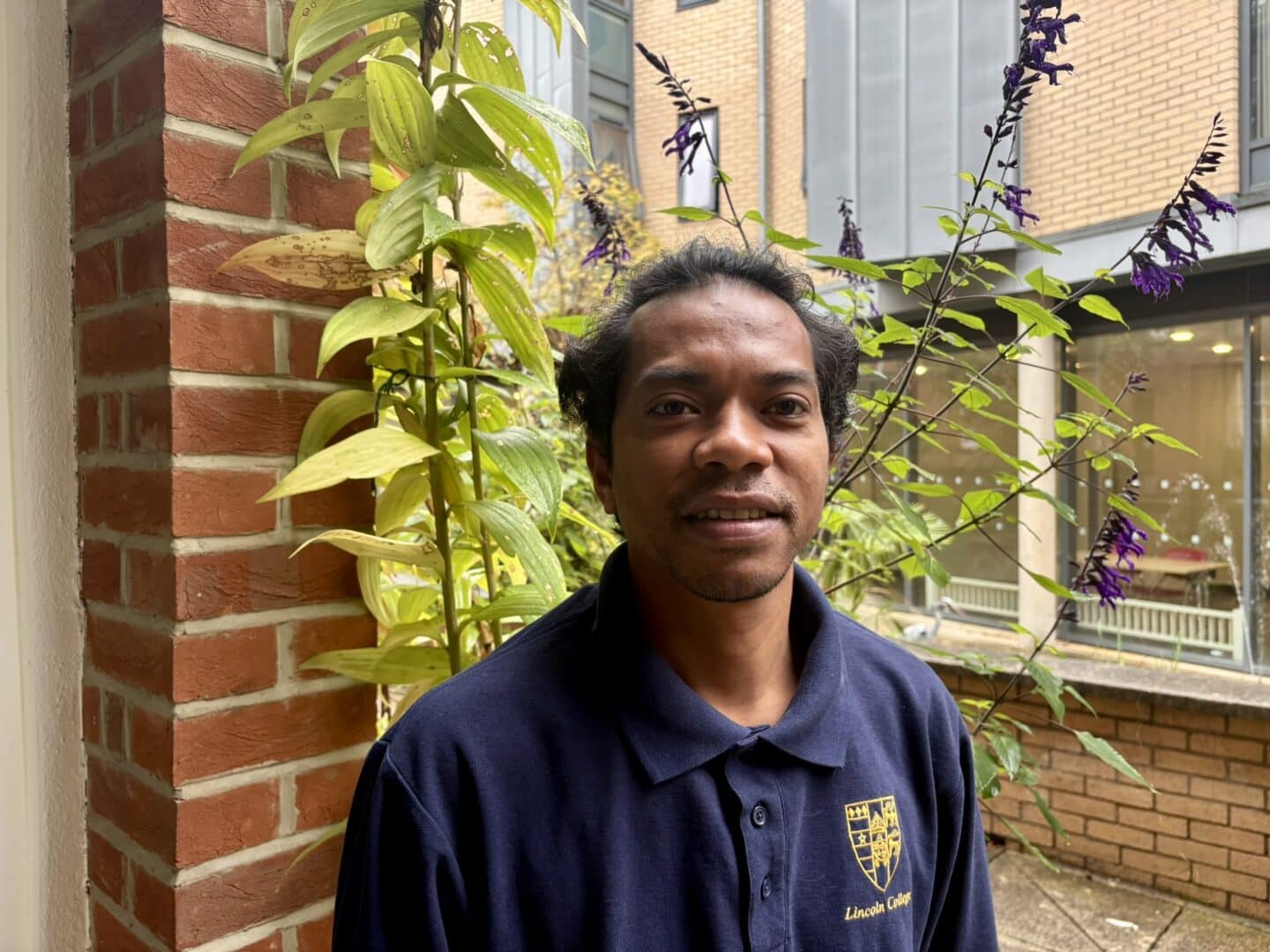 Cipriano Carralho, a Lincoln College scout, standing in a courtyard beside a brick pillar and flowering plants, with College buildings in the background.
