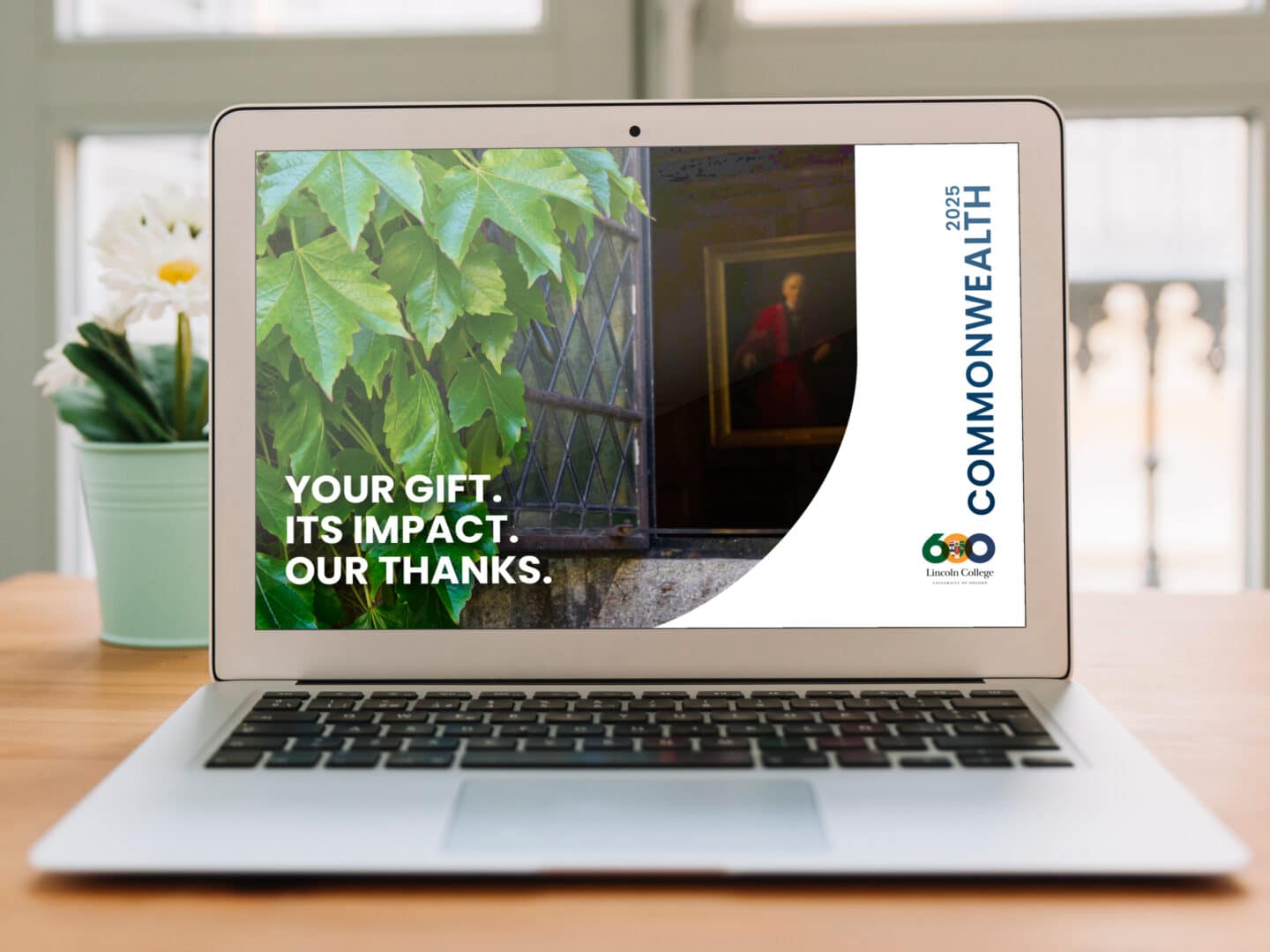 A laptop displaying the 2025 cover of Commonwealth, Lincoln College’s donor impact report. The design features ivy-covered stonework and a leaded window with a historic portrait inside. Bold text on the screen reads: "YOUR GIFT. ITS IMPACT. OUR THANKS." The Lincoln 600 logo and the word "COMMONWEALTH 2025" appear on the right-hand side of the screen. A green potted plant and daisy sit in the background on a wooden table.