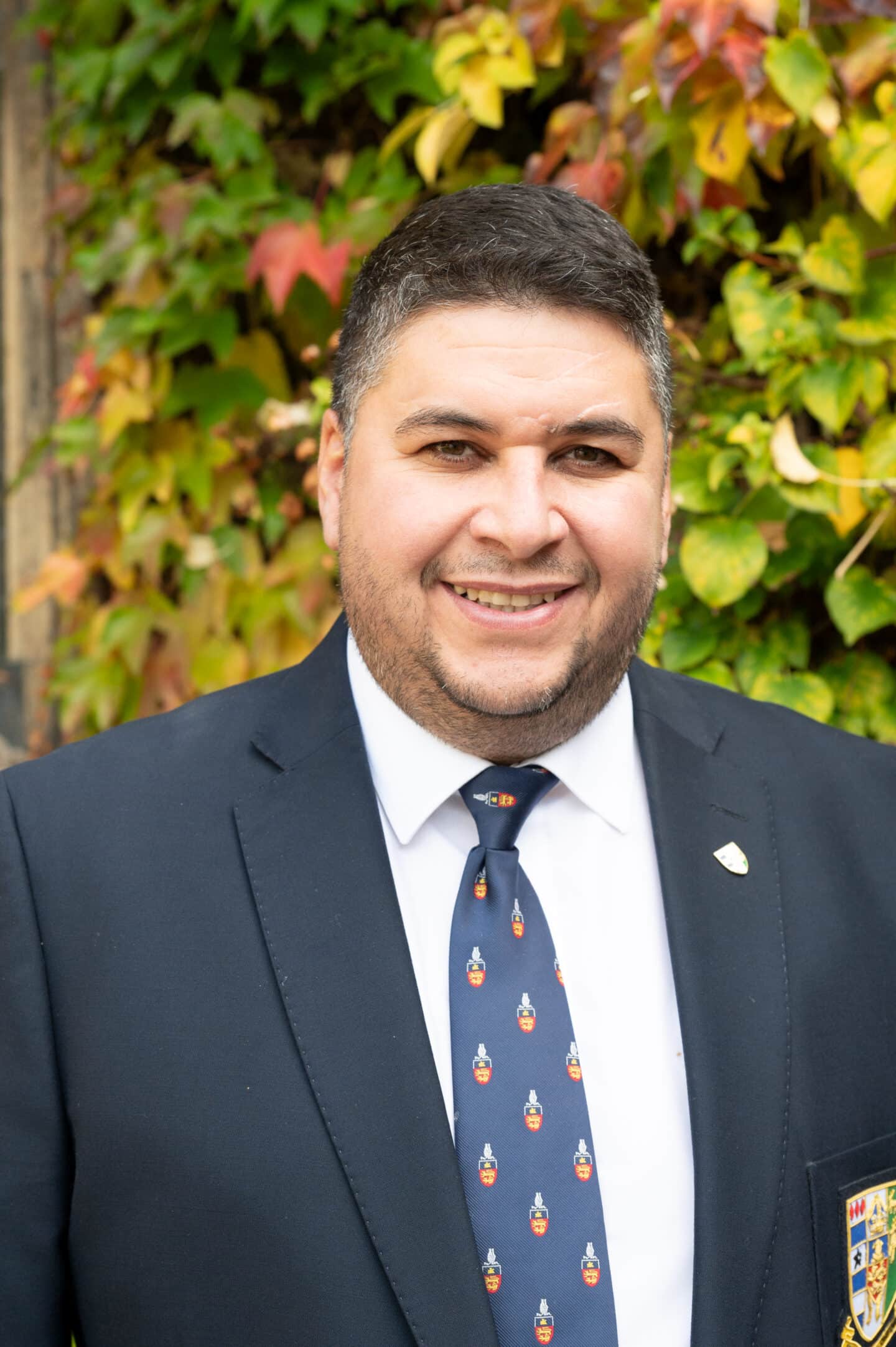 Cristiano da Silva standing in Front Quad at Lincoln College, Oxford, in a suit and College tie, smiling with ivy-covered wall behind.
