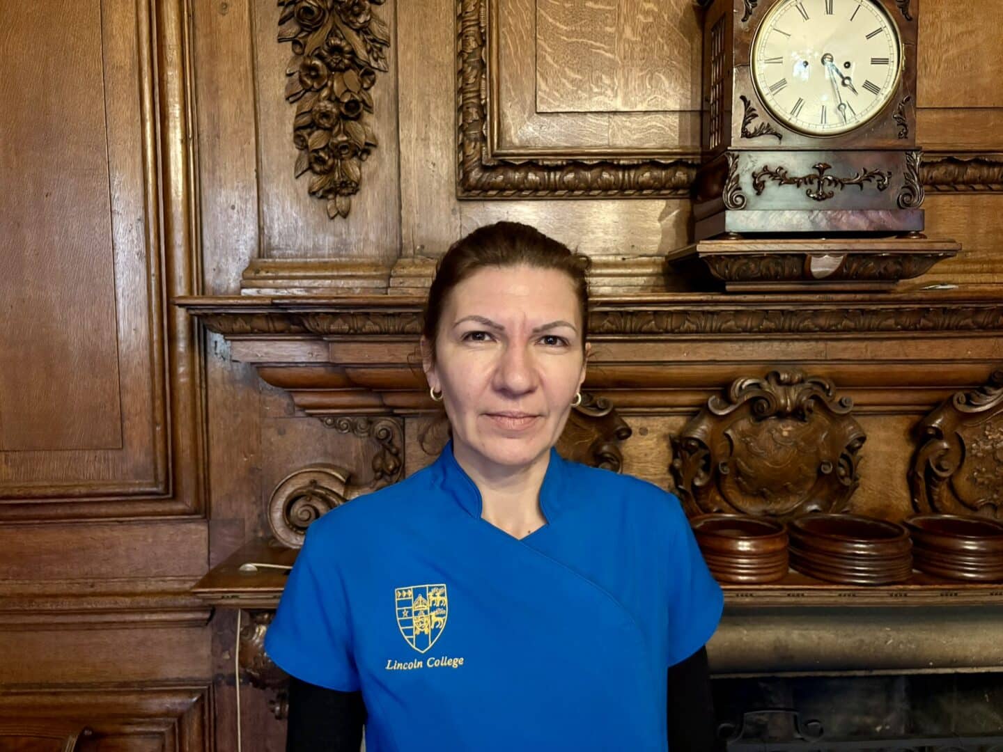 Cristina Vasiliu, Housekeeping Deputy Manager at Lincoln College, standing in front of an ornate wooden fireplace with carved detailing and a clock above, wearing a blue uniform with the College crest.