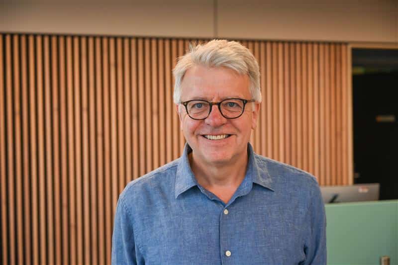 Stephan Chambers smiling indoors, wearing glasses and a blue shirt. He is standing in front of a modern wooden panelled wall, with soft lighting and a blurred reception desk in the background.