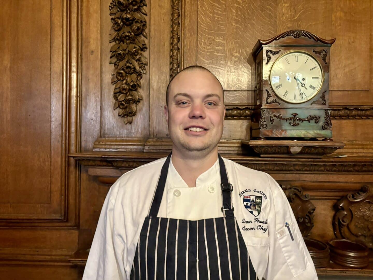 Dan Howells, Second Chef at Lincoln College, standing in front of an ornate wooden fireplace with carved detailing and a clock above, wearing a chef’s jacket and striped apron.