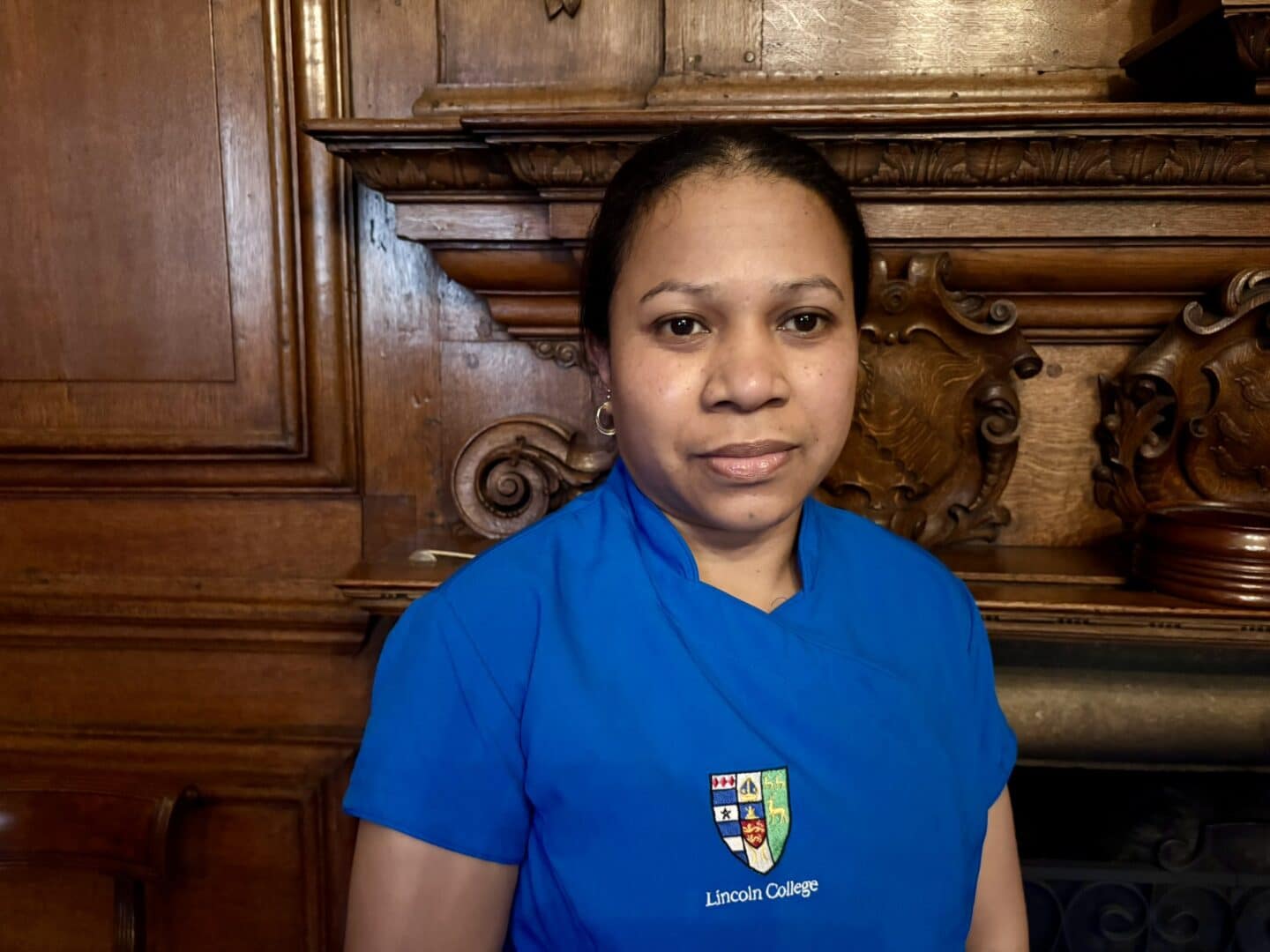Domingas Da Silva, a Lincoln College scout, standing in front of an ornate wooden fireplace with carved detailing, wearing a blue uniform with the College crest.