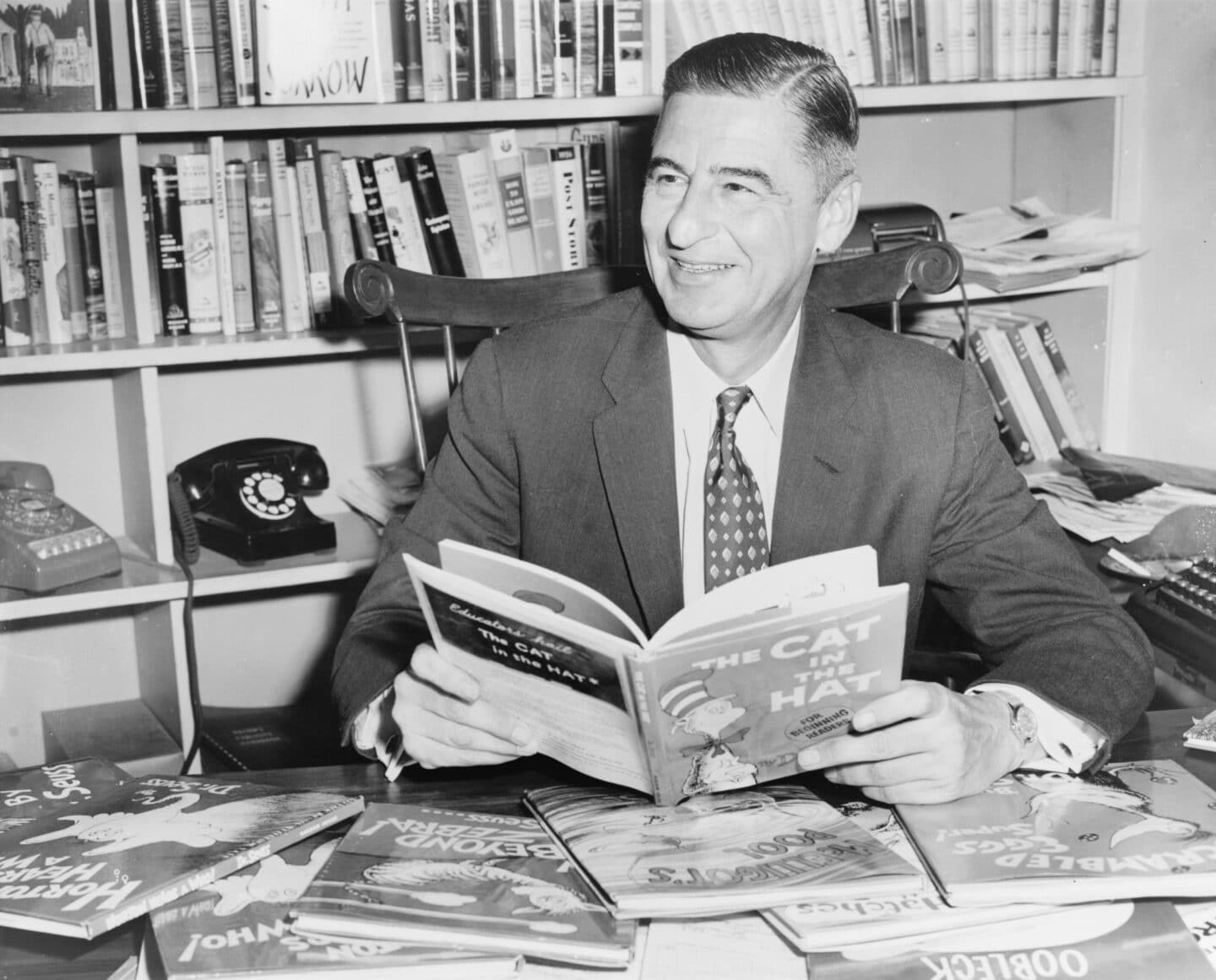 Author Theodor Seuss Geisel, known as Dr Seuss, sits at a desk smiling while holding an open copy of “The Cat in the Hat.” Several of his other illustrated books are spread out before him, and shelves of books fill the background.