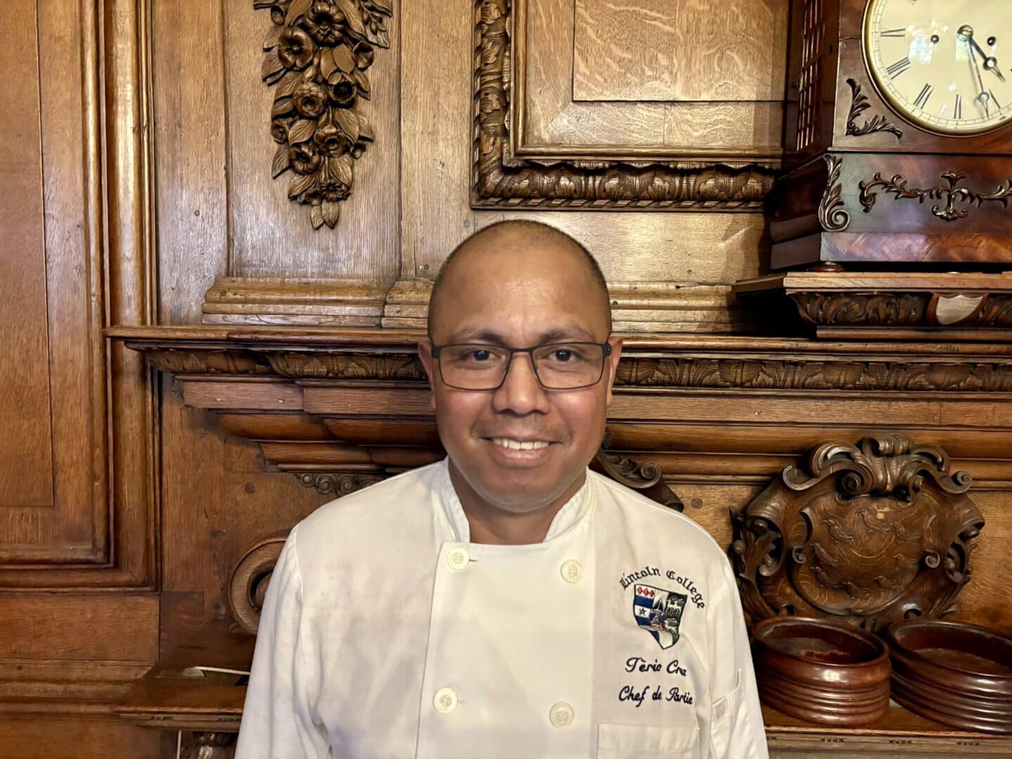 Eliterio Dos Santos Cruz, Chef de Partie at Lincoln College, standing in front of an ornate wooden fireplace with carved detailing and a clock above, wearing a chef’s jacket.