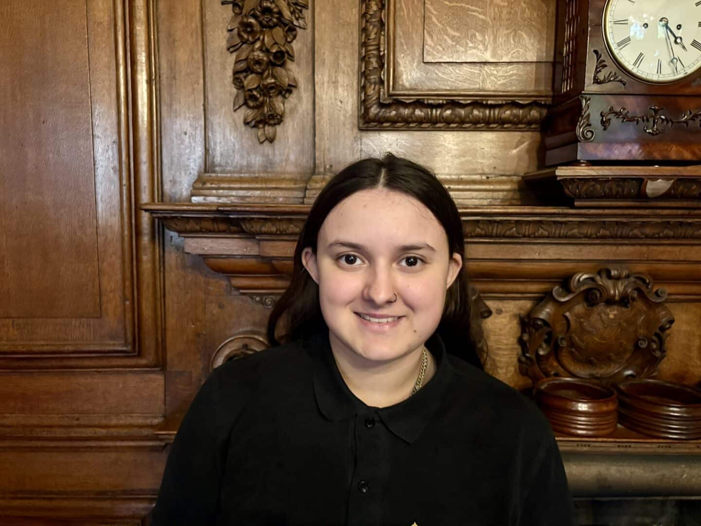 Lissa Radley, a Lincoln College scout, standing in front of an ornate wooden fireplace with carved detailing, wearing a black uniform with the College crest.