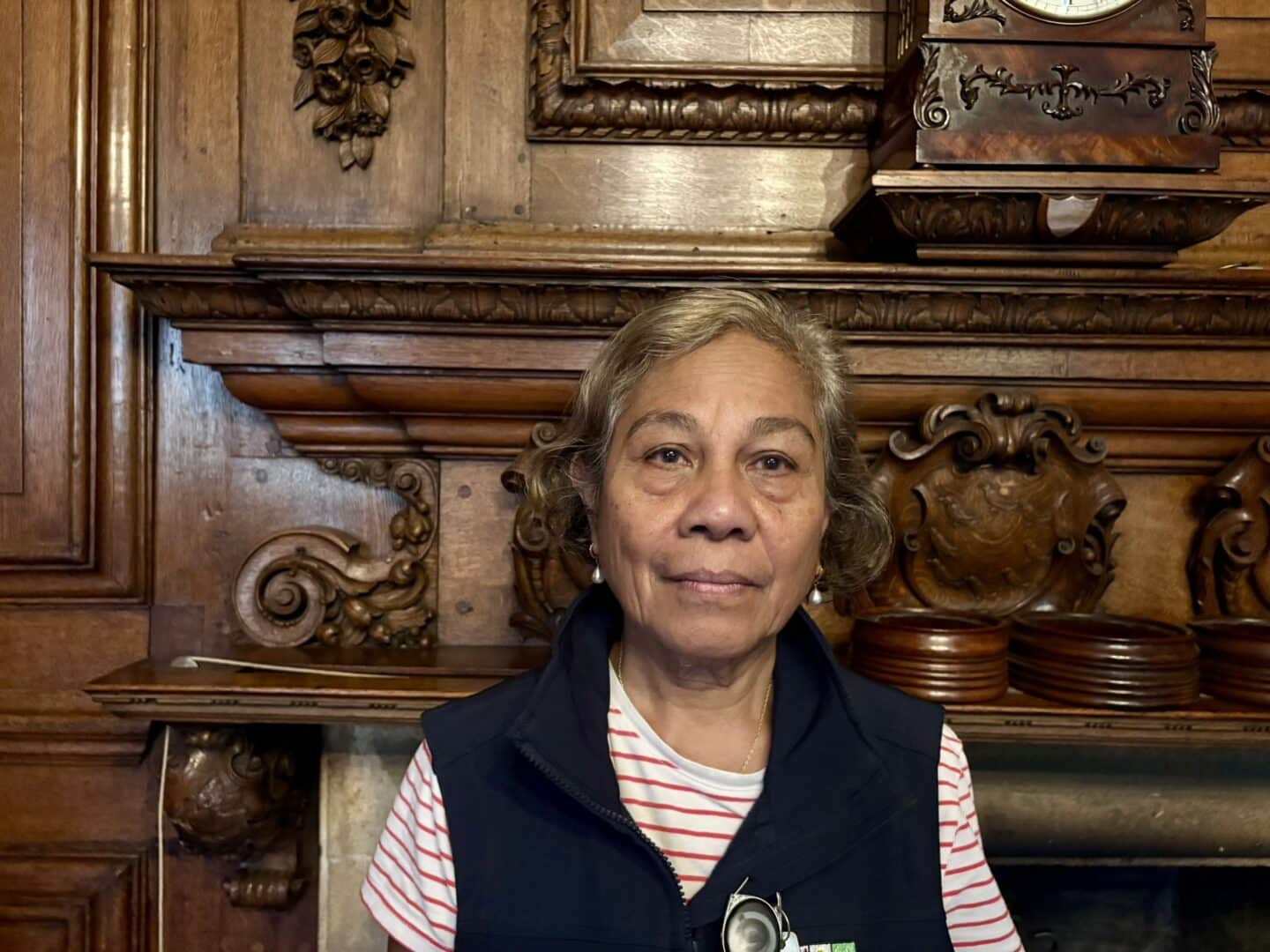 Ermelinda Ximenes, a Lincoln College scout, standing in front of an ornate wooden fireplace with carved detailing, wearing a striped top and dark vest with a College badge.