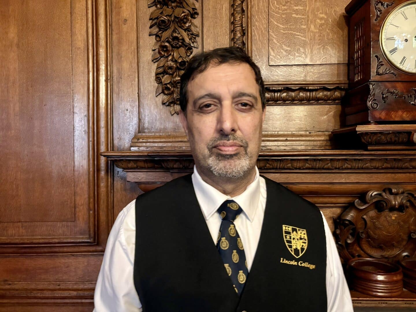Fida Hussain, Catering Supervisor at Lincoln College, standing in front of an ornate wooden fireplace with carved detailing and a clock above, wearing a white shirt, patterned tie, and black waistcoat with the College crest.