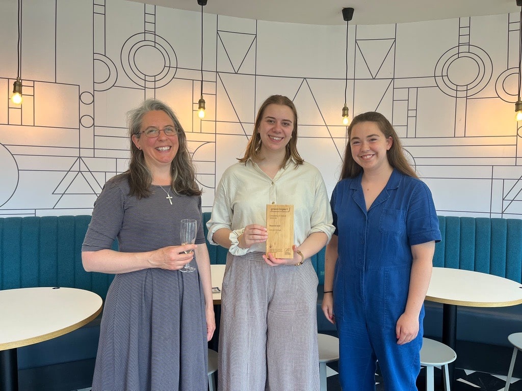 Lucy Matheson, Juliet Tye and Francesca Hammonds pose together indoors following the Green Impact awards. Juliet Tye stands in the centre holding Lincoln College’s wooden “Beyond Gold” award, with Lucy Matheson on the left and Fran Hammonds on the right. They are standing in front of a geometric patterned wall with pendant lights and teal banquette seating.