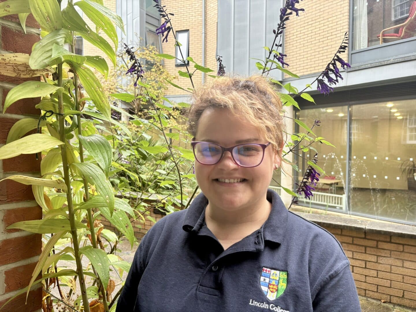 Hanna Gulba, a Lincoln College scout, standing in a courtyard beside a brick pillar and flowering plants, with College buildings in the background.