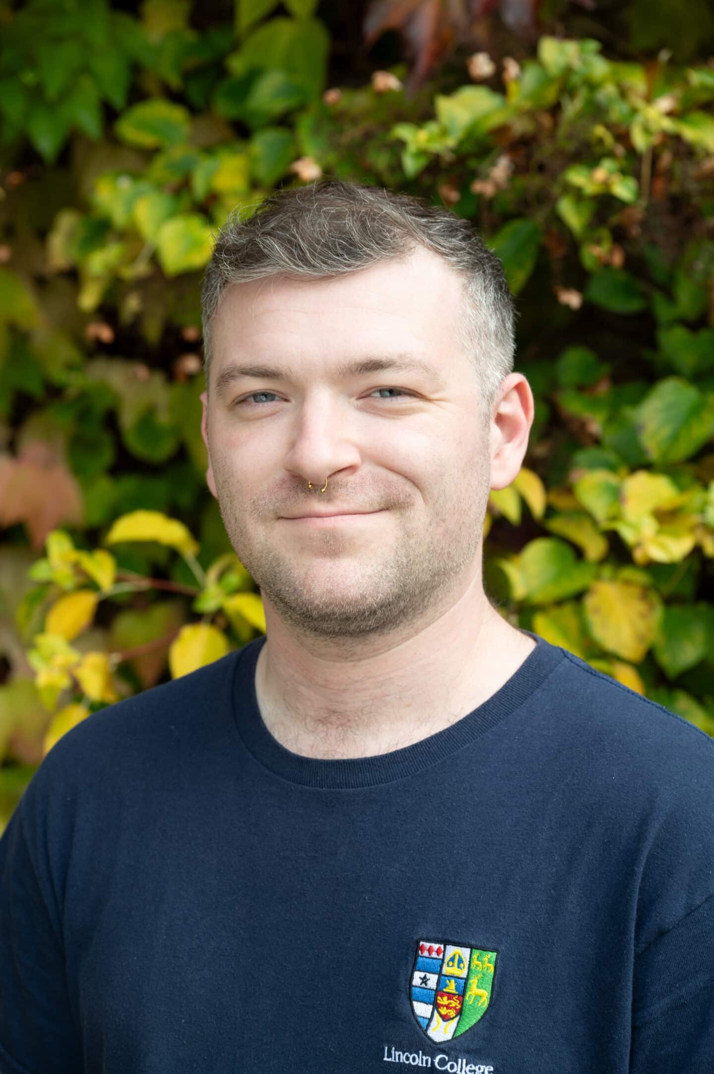 Bob Hembrow standing in Front Quad at Lincoln College, Oxford, wearing a navy Lincoln College T-shirt, with green and yellow ivy behind.