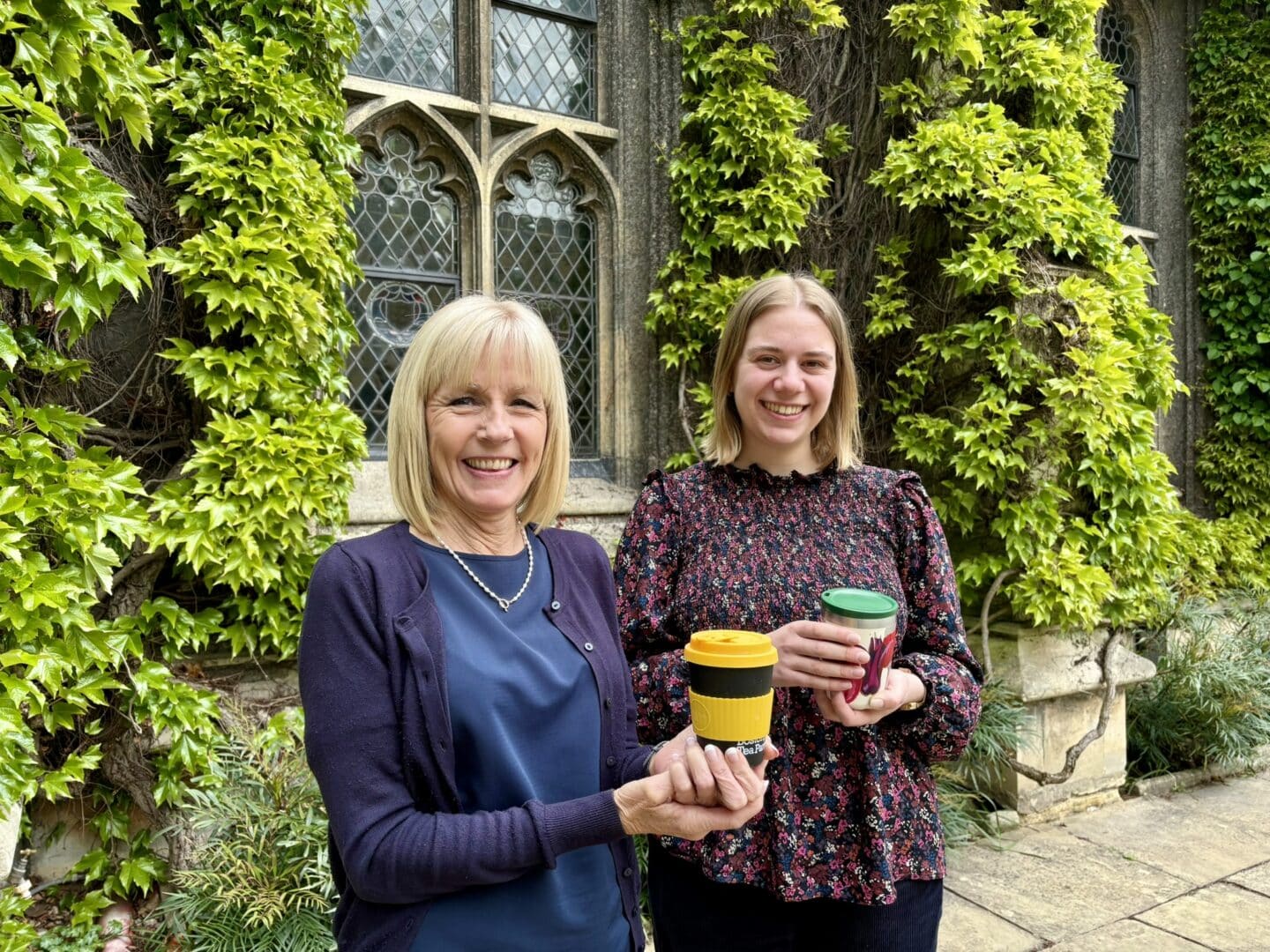 Susan Davison and Juliet Tye smiling and holding reusable coffee cups in front quad, standing in front of an ivy-covered stone Hall with ornate leaded glass windows.
