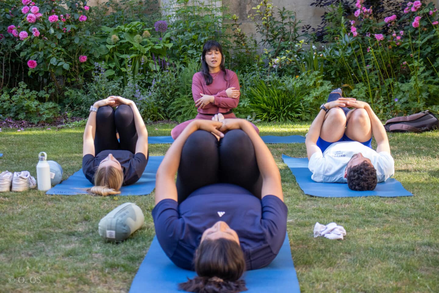 Micon leading a yoga session in the Rector’s Garden at Lincoln College, seated cross-legged in front of a group of students lying on mats in a gentle supine stretch. The garden is in full bloom behind them, with pink roses and lush greenery framing the tranquil scene.