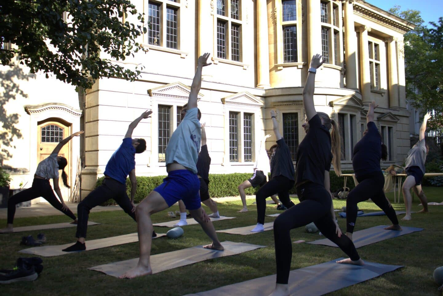 A group of Lincoln College students practising yoga in the Rector’s Garden, framed by the historic architecture of Lincoln College in the background. The students are in warrior pose, stretching on yoga mats in the afternoon light.