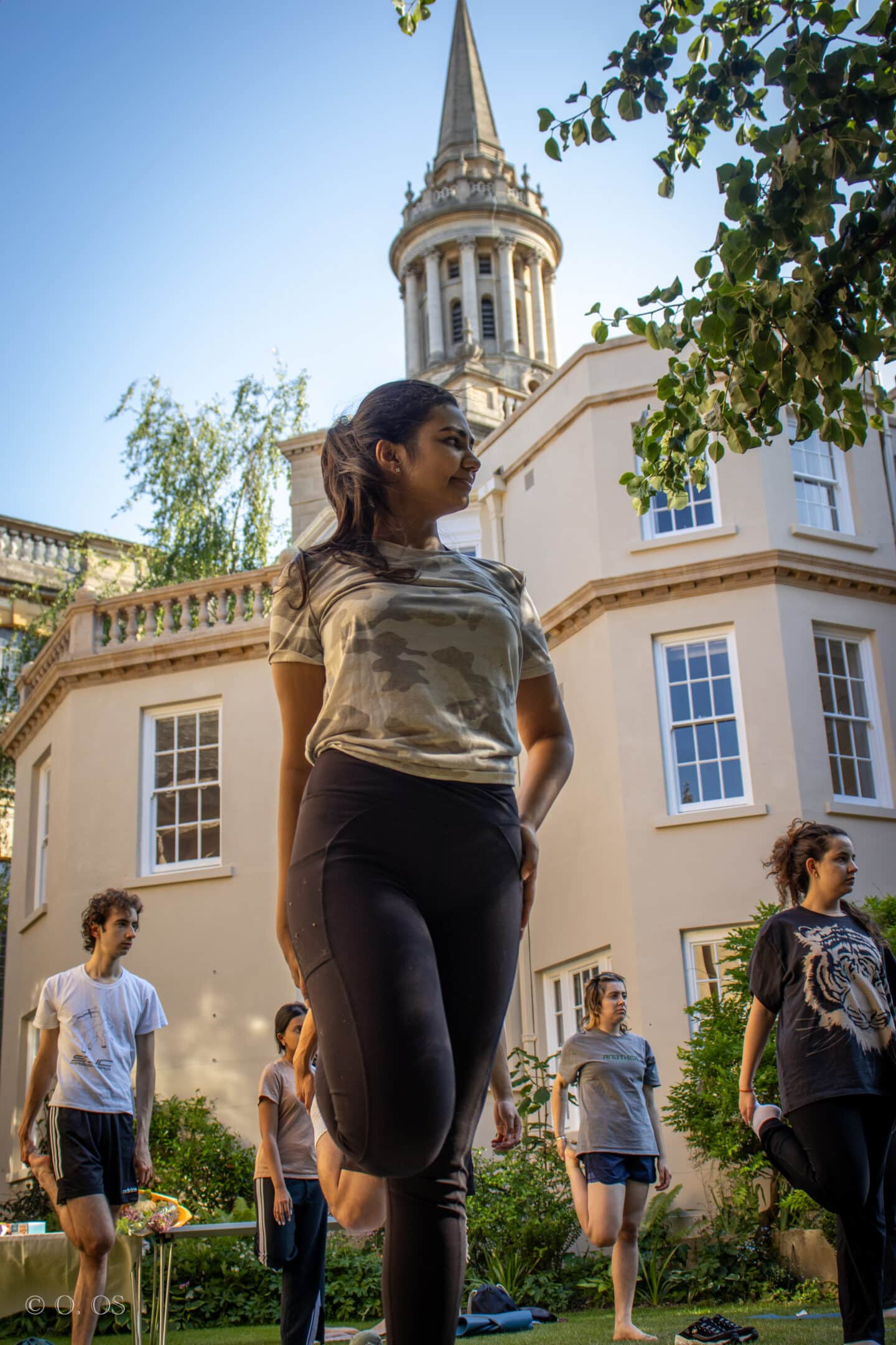 A Lincoln College student stands in a balancing pose during a yoga session in the Rector’s Garden, with the iconic spire of Lincoln College rising behind her. Other students are visible in the background, mid-stretch, in the warm glow of a sunny afternoon.