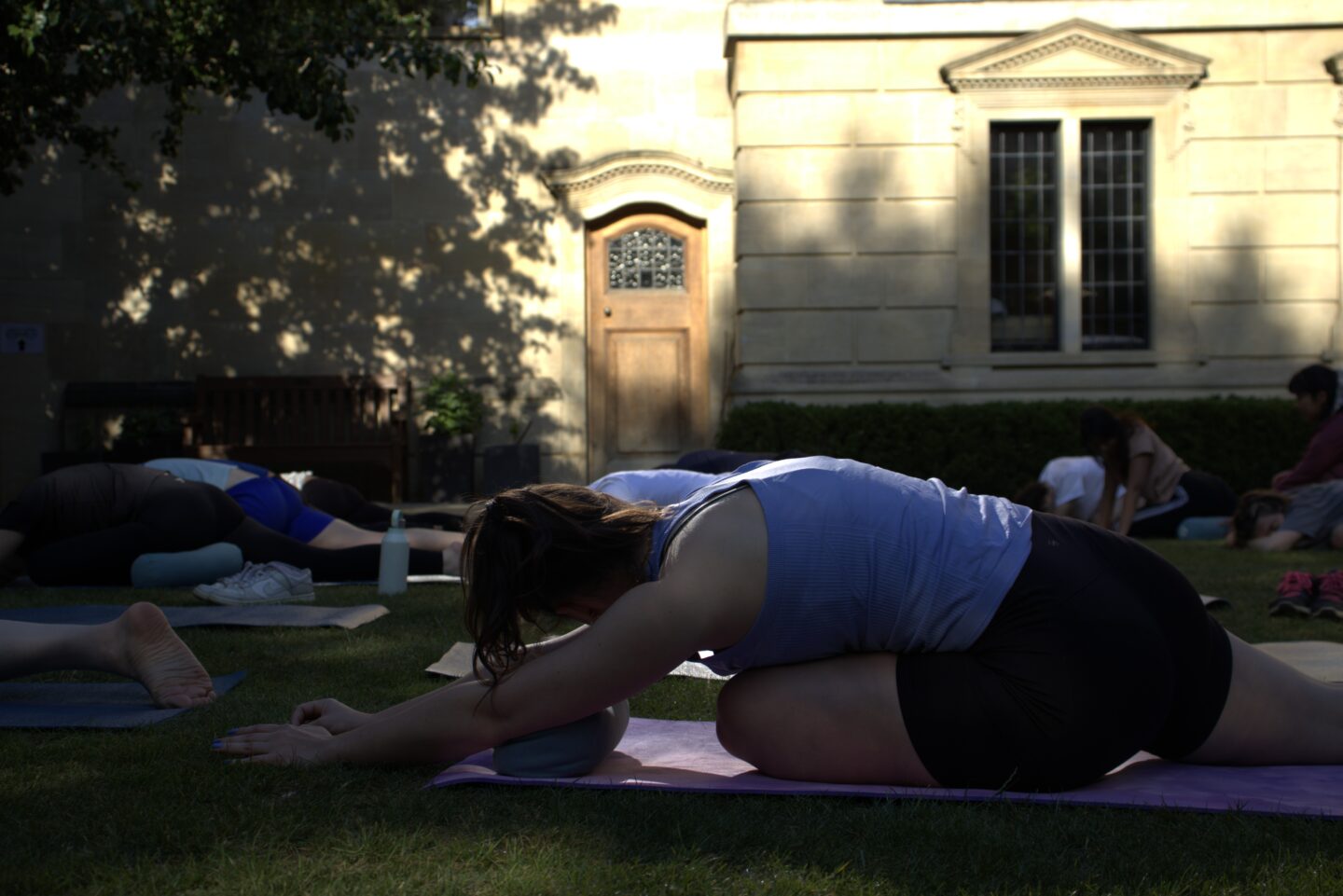 Lincoln College students taking part in a yoga session in the Rector’s Garden, with participants in a seated forward fold pose using bolsters for support. The warm stone walls and arched door of the College provide a peaceful backdrop in the afternoon shade.