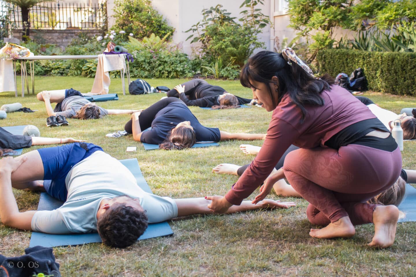 Micon guiding a Lincoln College student through a gentle spinal twist during an outdoor yoga session in the Rector’s Garden. Other students lie on mats around the lawn, surrounded by summer greenery and relaxed atmosphere.