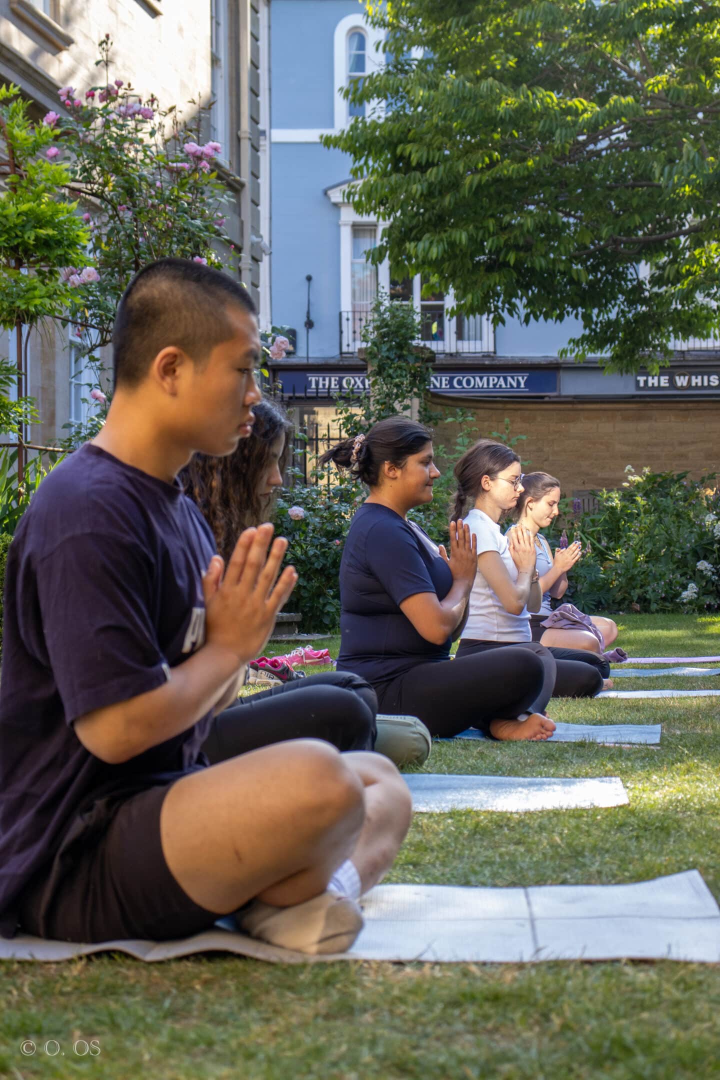 Lincoln College students seated cross-legged on yoga mats in the Rector’s Garden, hands in prayer position during a moment of quiet reflection. A backdrop of roses, greenery, and the Oxford Wine Company building beyond the College walls adds a serene and distinctly Oxford touch.