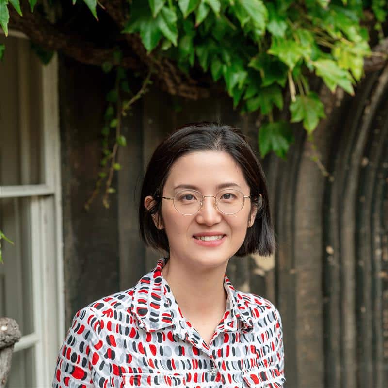 Tiffany Luo standing in front of a stone archway at Lincoln College, Oxford, smiling at the camera. She is wearing glasses and a patterned shirt with red, black and grey details.