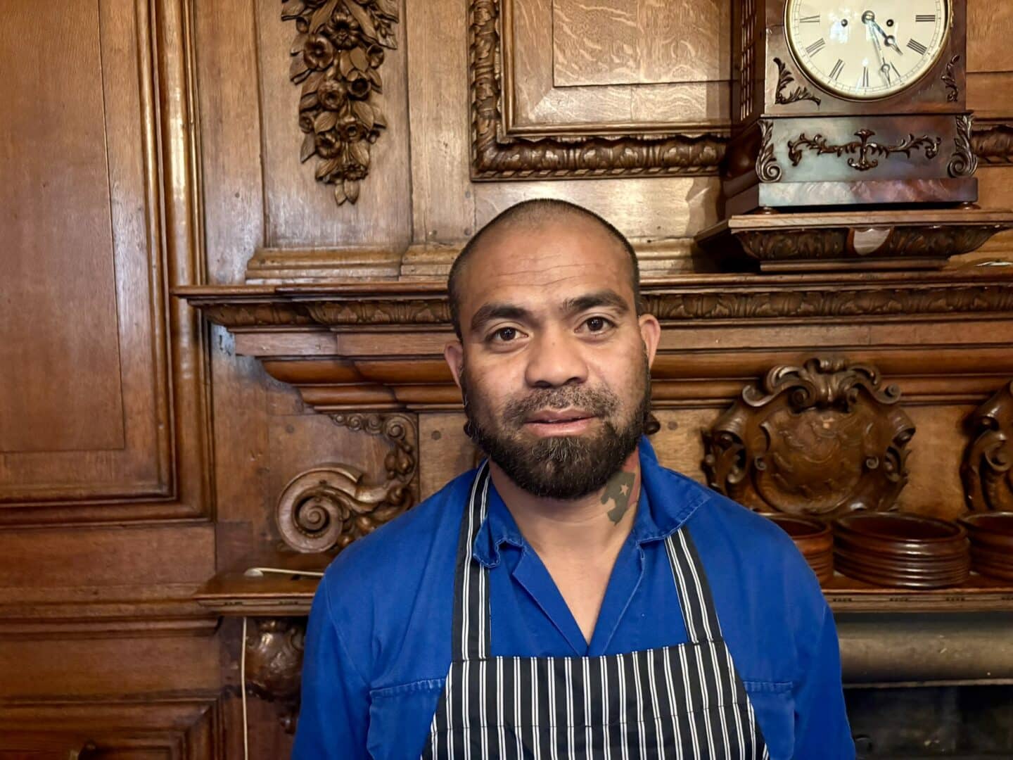 Inocencio Da Costa, a Lincoln College kitchen porter, standing in front of an ornate wooden fireplace with carved detailing, wearing a blue uniform and striped apron.