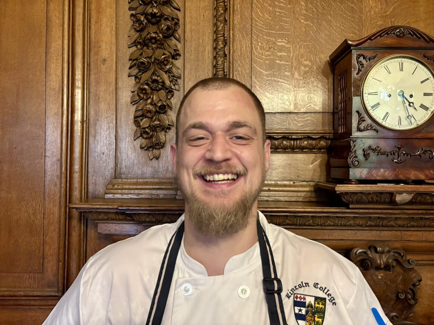 Jake Franklin, Chef de Partie at Lincoln College, standing in front of an ornate wooden fireplace with carved detailing and a clock above, wearing a white chef’s jacket with the College crest and a black apron.