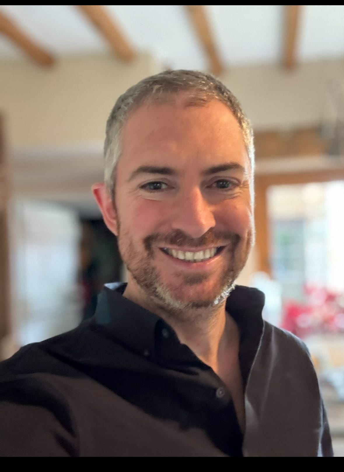 Jerome Glass smiling indoors, wearing a dark shirt. The background shows soft lighting, wooden ceiling beams, and blurred furnishings, creating a warm, informal setting.