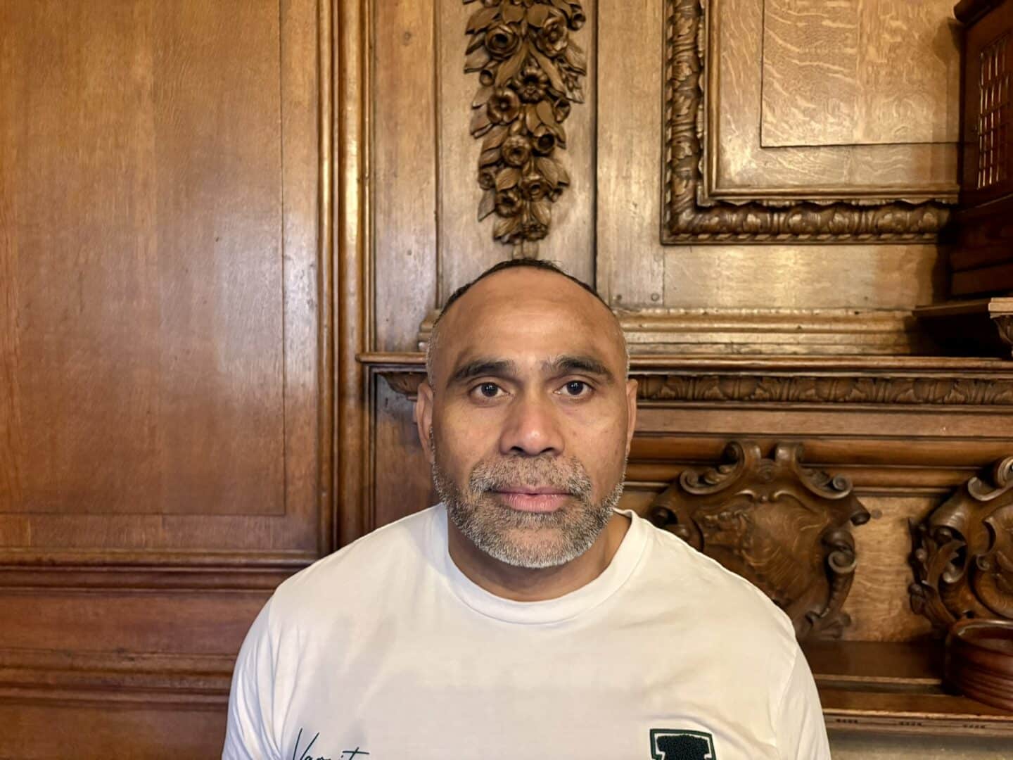 Joaquim De Jesus Antunes, a Lincoln College kitchen porter, standing in front of an ornate wooden fireplace with carved detailing, wearing a white T-shirt.