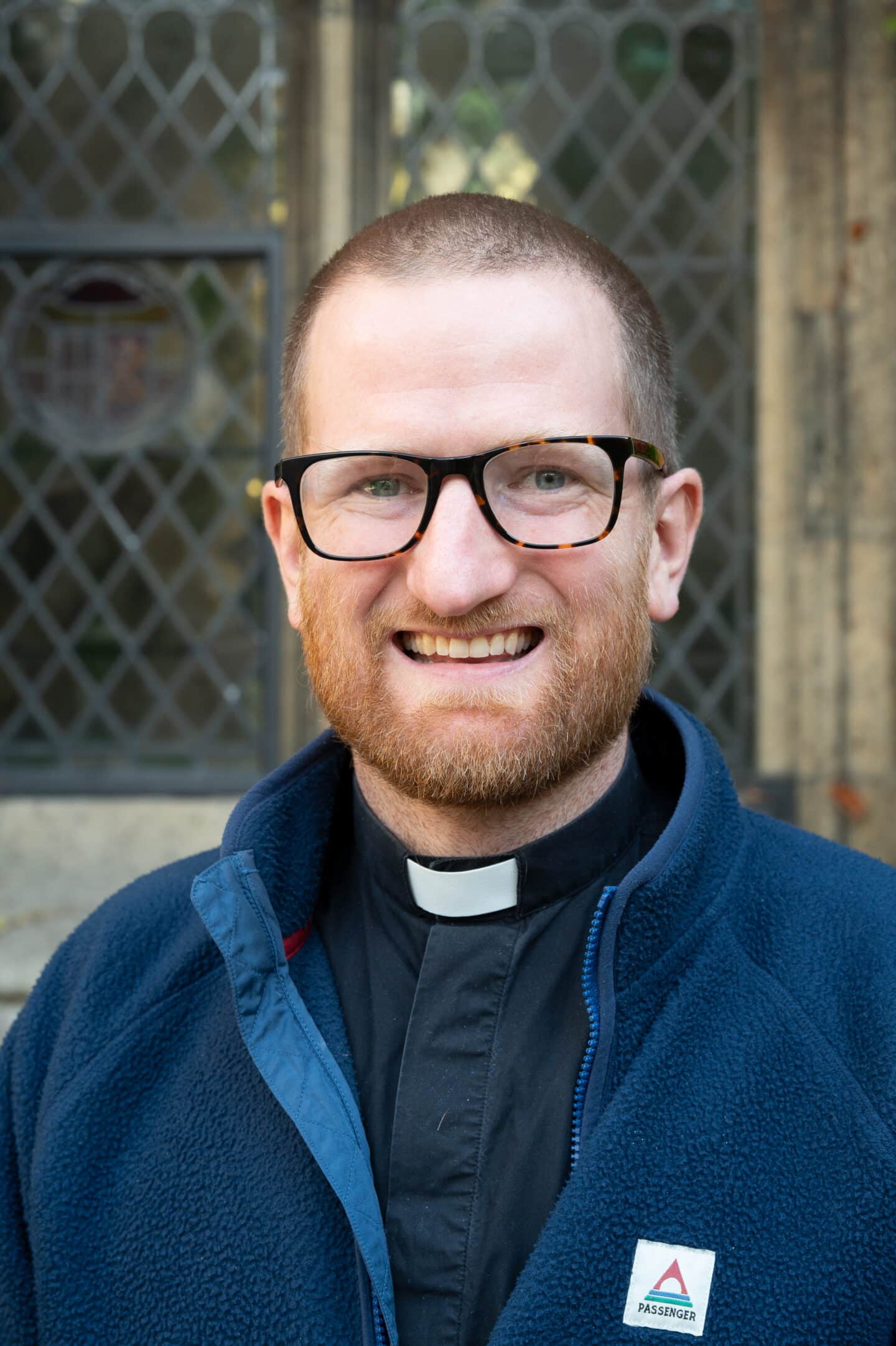 Revd Jonny Torrance standing in Front Quad at Lincoln College, Oxford, wearing clerical collar and navy fleece, with leaded window and ivy behind.
