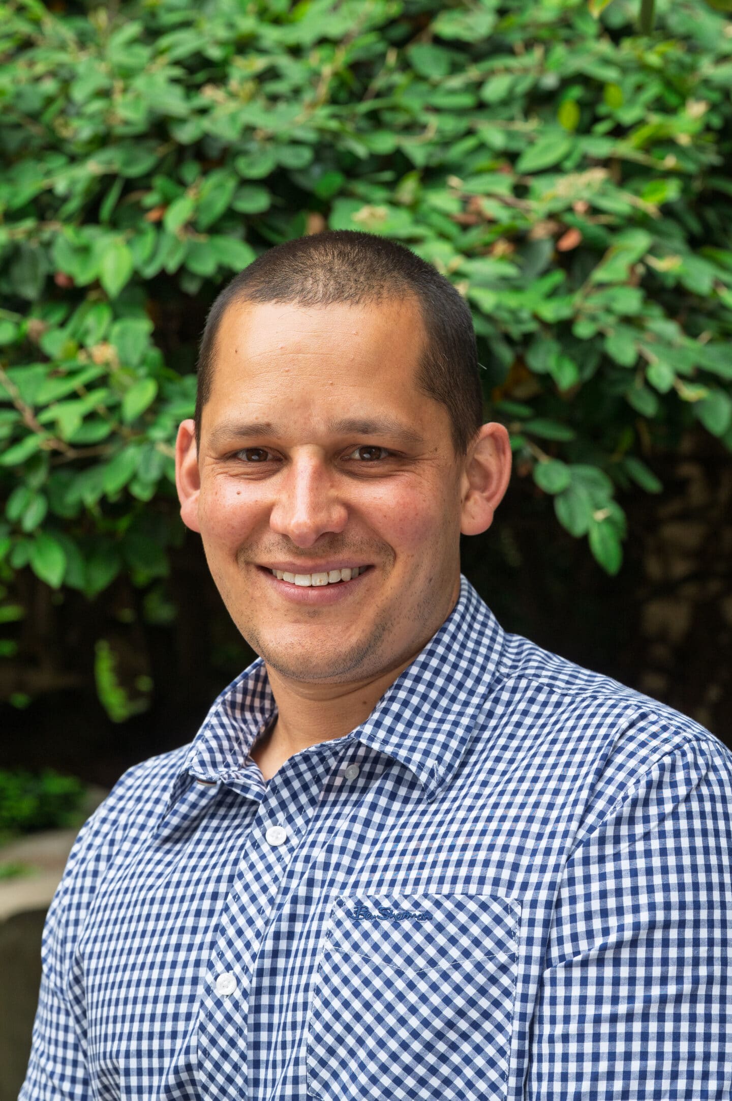Portrait of Kiran Isaac wearing blue and white checked shirt against greenery
