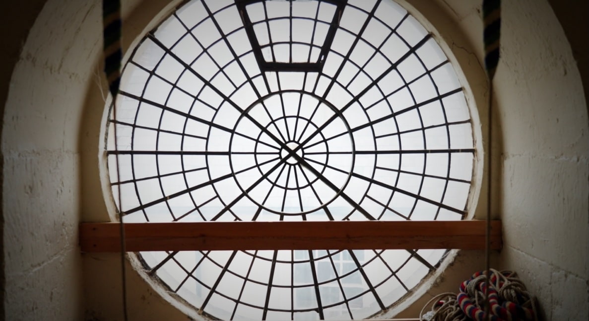 Interior view of a circular window featuring a radial iron grid, framed by thick stone walls with soft natural light coming through.