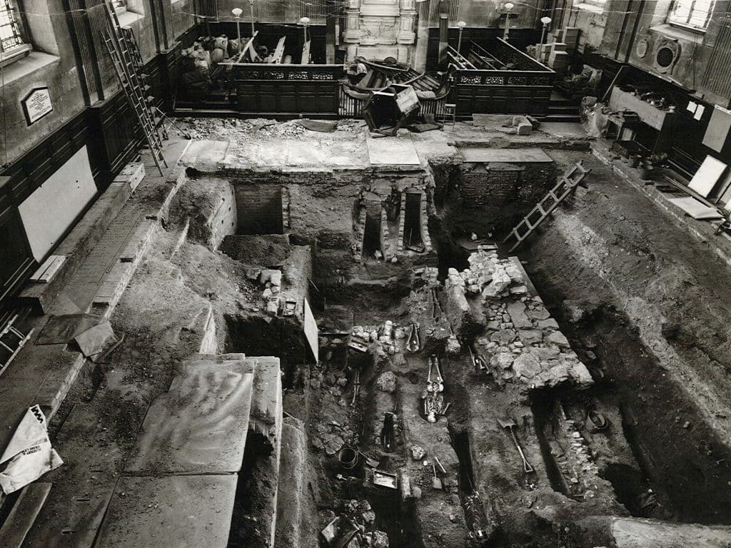 Black-and-white photograph of an excavation inside the Library building, revealing skeletons, stone walls, and graves beneath the floorboards.