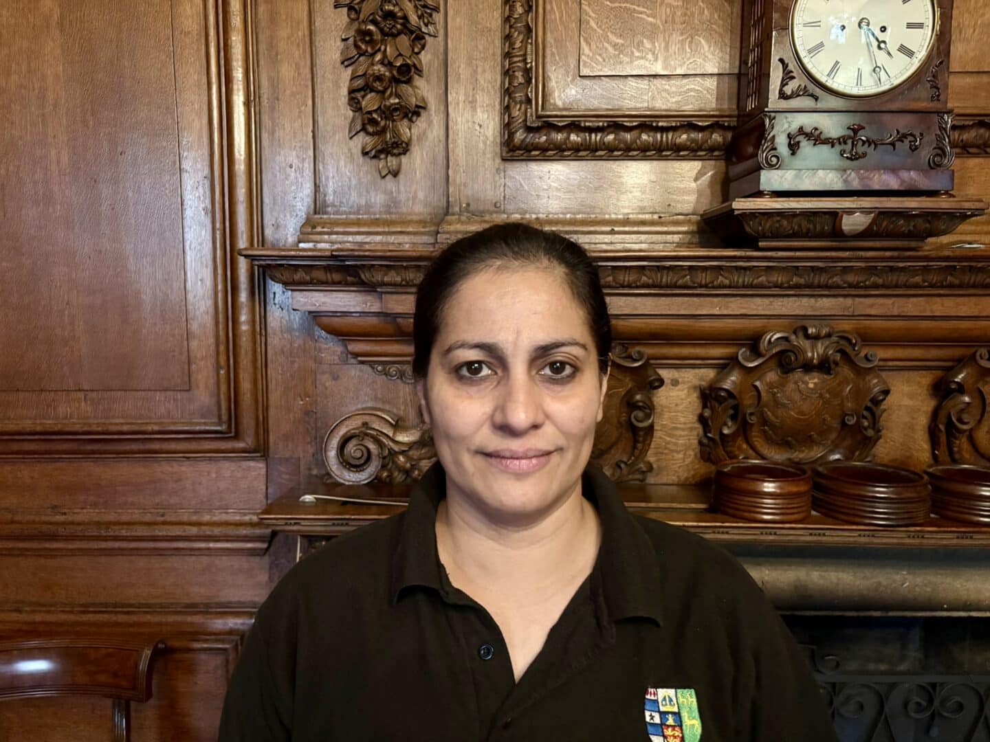 Ligia Duarte, Catering Assistant at Lincoln College, standing in front of an ornate wooden fireplace with carved detailing and a clock above, wearing a black polo shirt with the College crest.