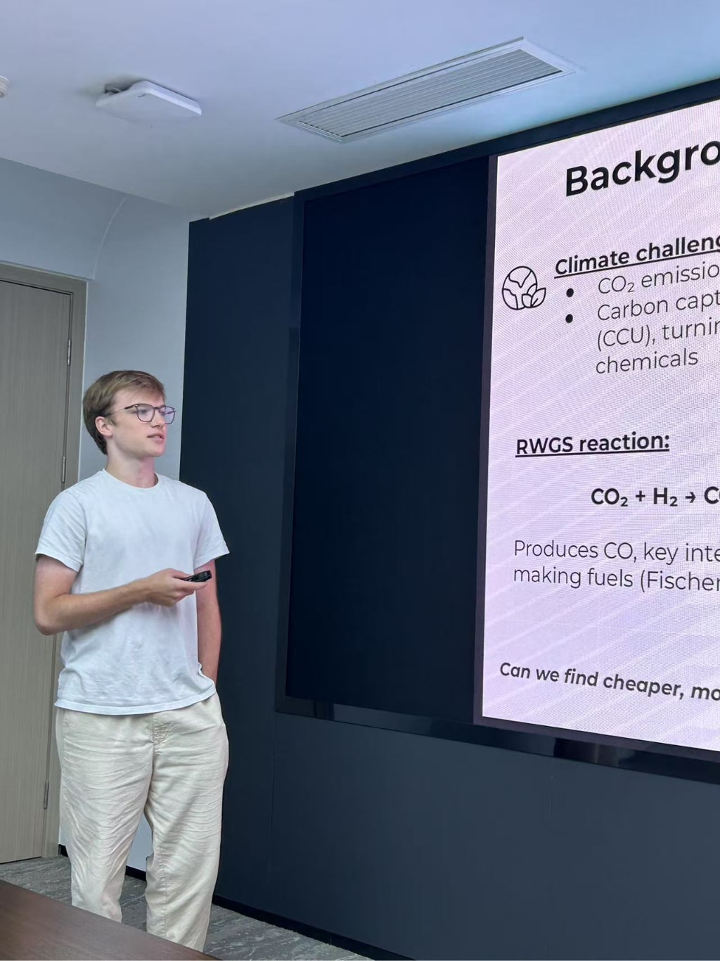 Luca Viviano delivering a presentation in a conference room, standing beside a large screen displaying research findings on climate challenges and carbon capture reactions.