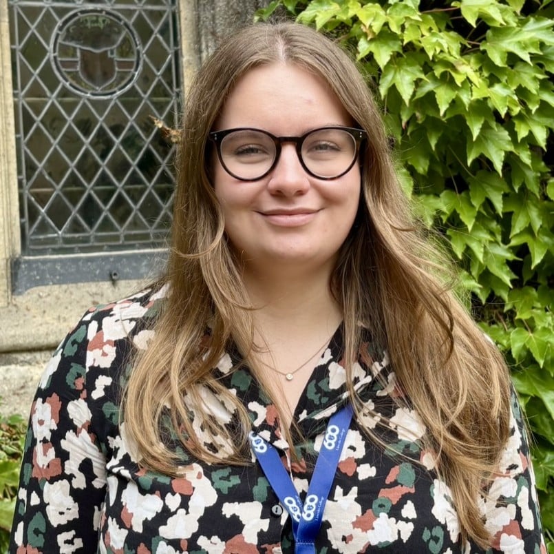 A head and shoulders photograph of Lucy Whiting. She has long, light-brown hair, is wearing black-rimmed glasses and a patterned blouse, and is smiling at the camera. She is standing outside in front of a stone building in Front Quad with ivy on the wall.