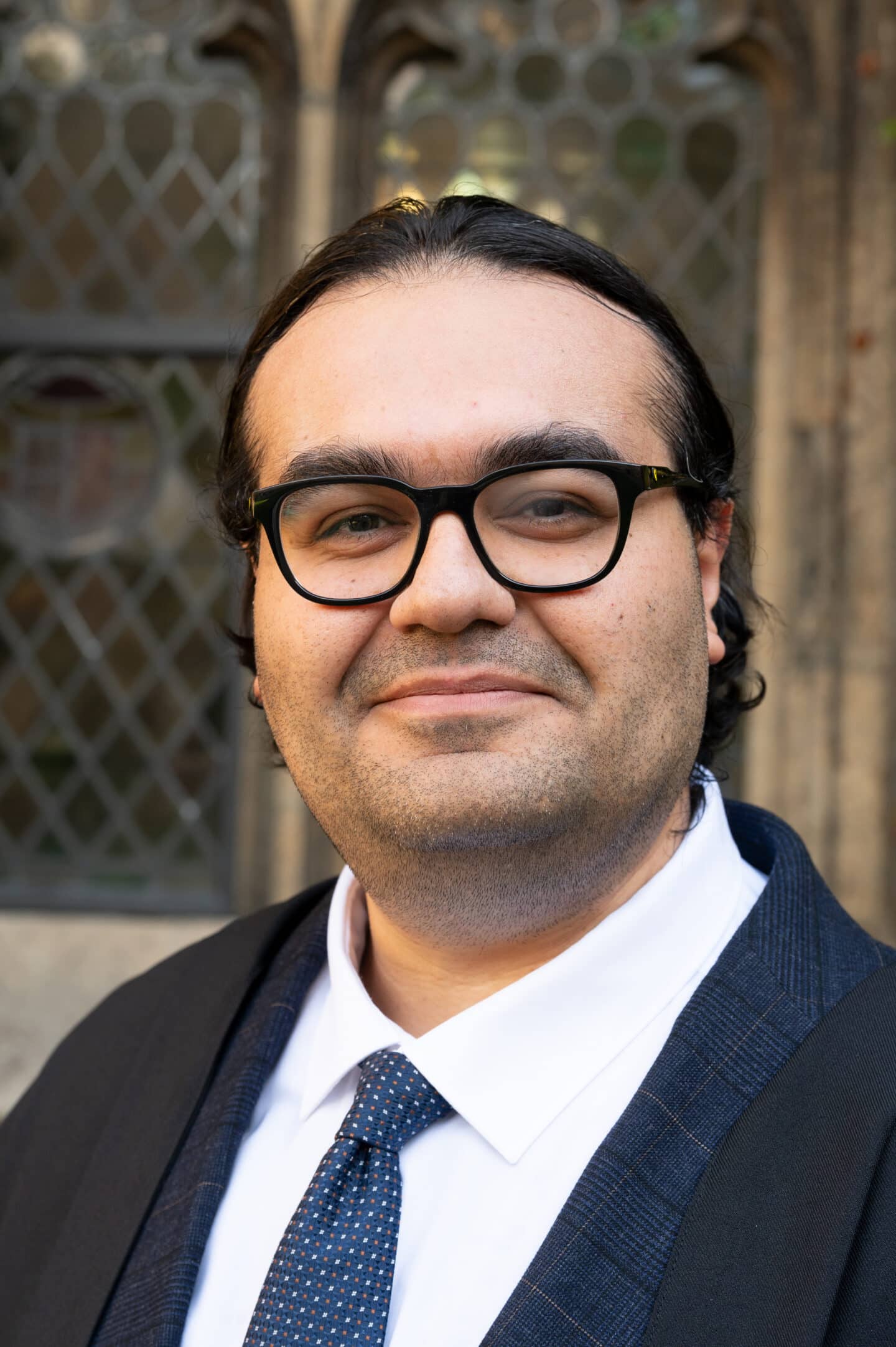 Mahan Ghafari standing in Front Quad at Lincoln College, Oxford, wearing a dark suit and tie, with a leaded window and stonework behind.