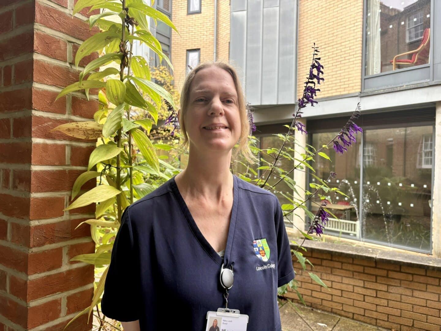 Mary Louth, a Lincoln College scout, standing in a courtyard with plants and brick buildings in the background.